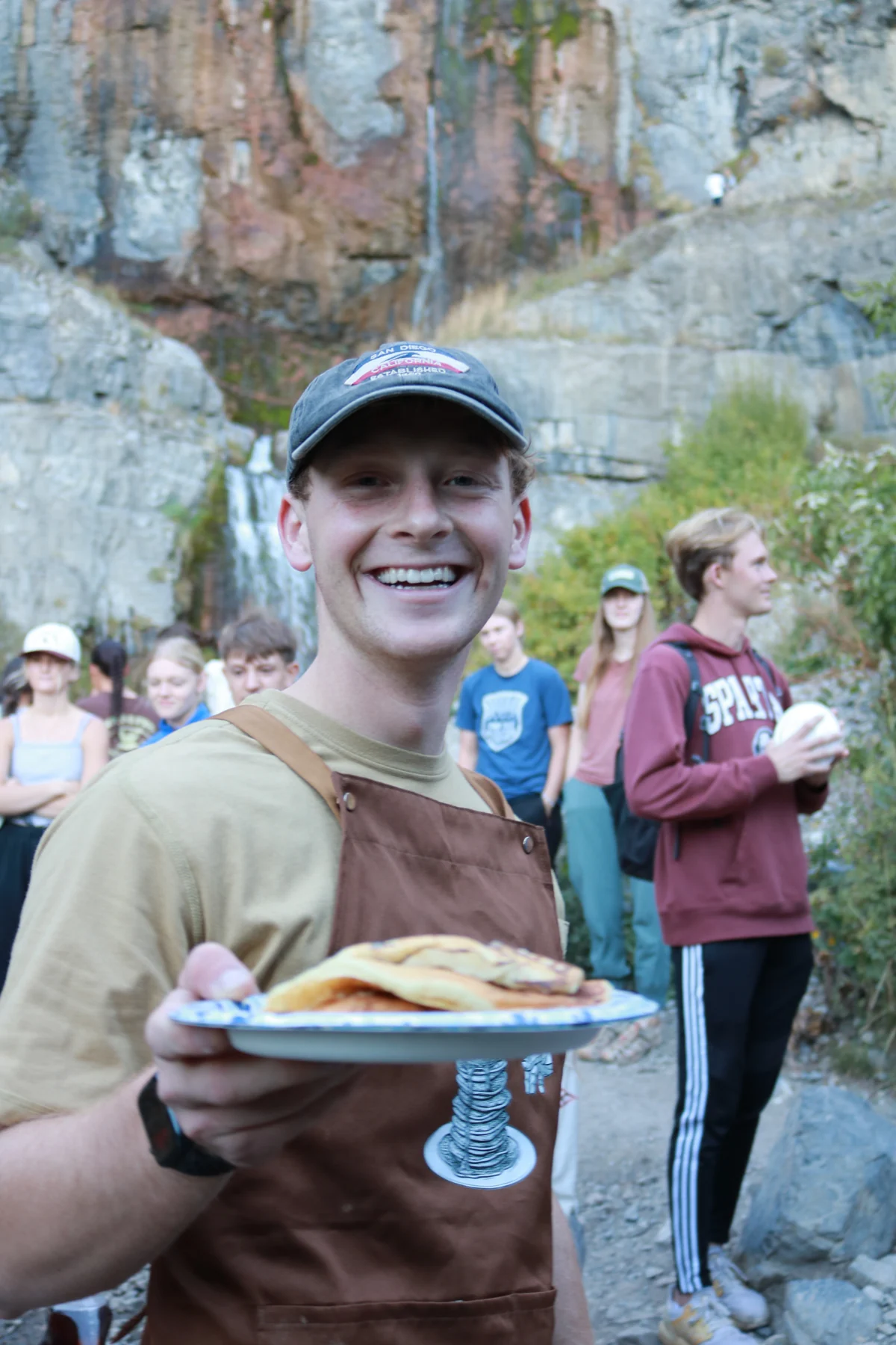 A smiling Pancake Boy wearing the custom brown apron and holding a plate of pancakes